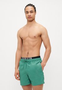 Pantaloni scurți de înot verzi cu talie ajustabilă și buzunare laterale, având un elastic negru cu brandul 'CALVIN KLEIN'. Textură netedă.