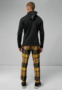 Felpa nera con cappuccio, abbinata a pantaloni a quadri gialli, neri e verdi. Le sneakers completano l'outfit.