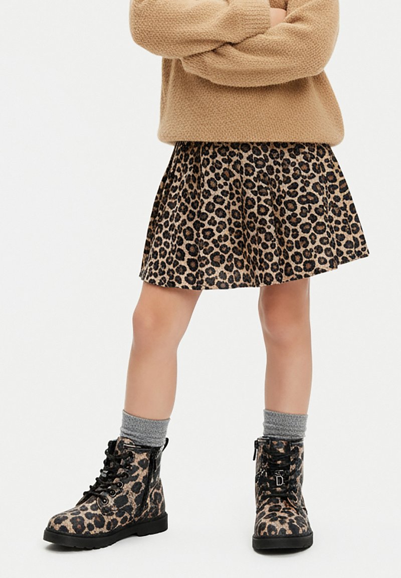 Kind trägt einen Leopardenmuster-Rock und passende Schnürstiefel, graue Socken und einen beigen Strickpullover, die Arme verschränkt.