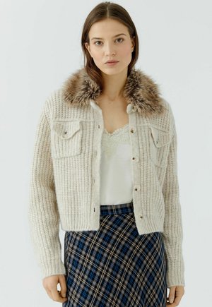 Junge Frau trägt eine cremefarbene Strickjacke mit Fellkragen über einem weißen Spitzenoberteil und einem blau karierten Rock, steht vor einem schlichten Hintergrund.