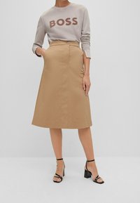 Beige A-linjekjol med knappstängning framtill och sidofickor, kombinerad med en grå sweatshirt med ordet "BOSS" i brunt. Svarta klacksandaler.