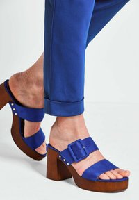 Blauwe platform sandalen met een brede band, gespsluiting en houten hak. De textuur is glad met gouden studs op de band.