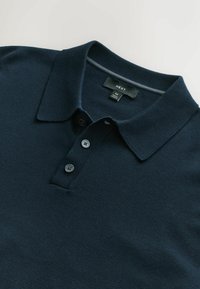 Tumed polo-särk pehmest kangast, millel on klassikaline krae, kolme nööbiga nööbirea ning soonikuga mansetid. Brändi silt on nähtav.