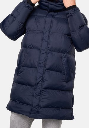 Osoba nosząca granatową puchową kurtkę z kapturem, poprawiająca kołnierz, stojąca na tle jednolitego jasnego tła.