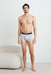 Boxers grises avec une ceinture élastique bleu marine arborant le logo "LACOSTE", ornés de petits points bleus.