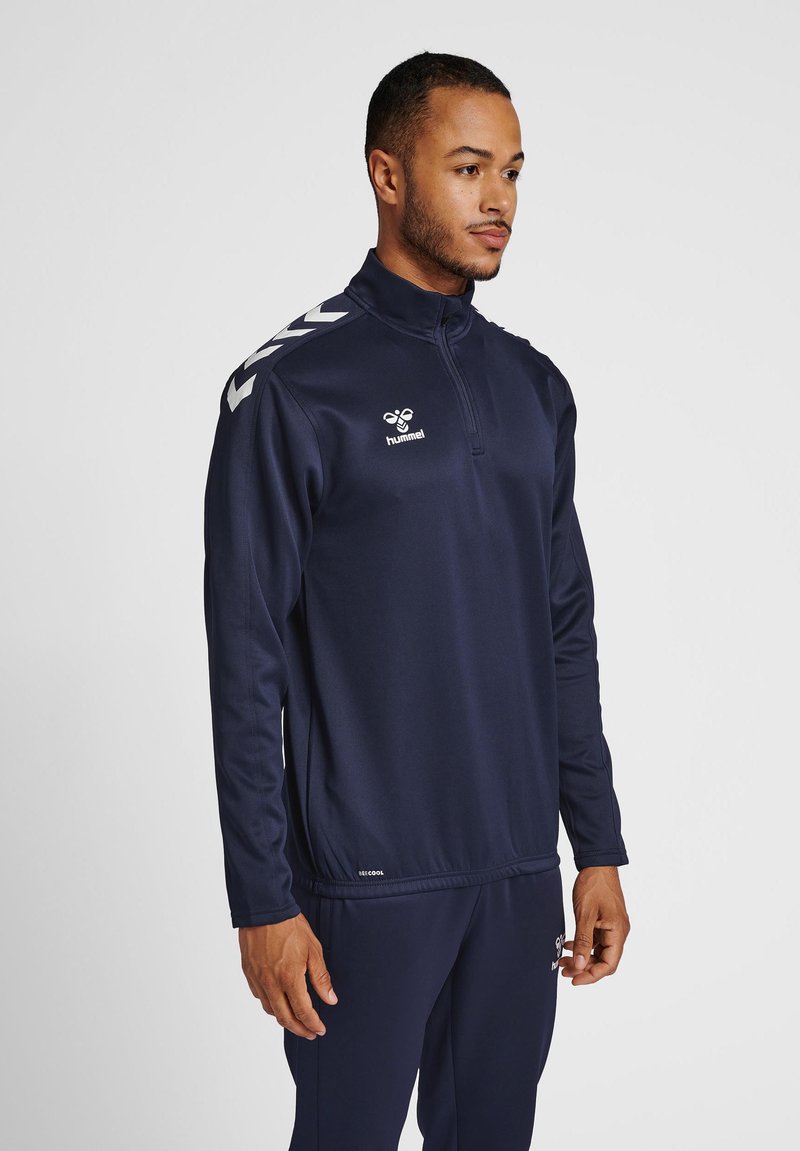 Mann, der eine marineblaue Sportjacke mit Viertelreißverschluss und passende Hose trägt, steht da und schaut seitwärts vor einem einfarbigen Hintergrund.