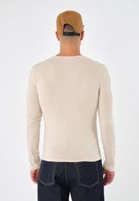 Beige långärmad skjorta med rundad halsringning, mjuk textur och figurnära design, kombinerad med mörkblå denimbyxor och en brun keps.