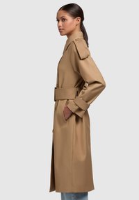 Dlouhý béžový trench coat s páskem v oblasti pasu, bočními kapsami a ramenními vycpávkami. Hladká látka s knoflíkovou légou a klasickým límcem.