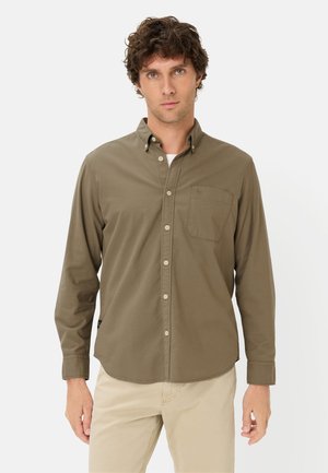 Young man wearing olive green button-up shirt and beige pants, standing against a plain light background, facing forward.