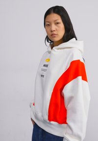 Hoodie blanc avec accent orange, coupe oversize, présentant un logo et un texte à l'avant. Matière douce, capuche avec cordon de serrage et poignets côtelés.