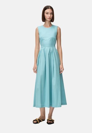 Donna in piedi che indossa un vestito midi azzurro chiaro senza maniche e sandali neri con punta aperta e dettagli in corda beige su sfondo bianco.