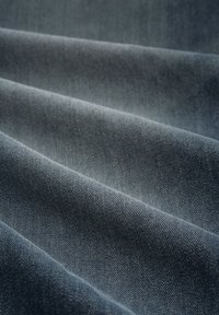 Tumši denima audums, kas parādīts salocītās kārtās, ar gludu tekstūru un maigu mirdzumu, ar redzamām šuvju detaļām tuvplānā.