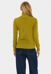 Olivno zelena turtleneck pulover s prilagojenim dizajnom, rebrastimi manšetami in spodnjim robom, v kombinaciji s temno modrimi širokimi hlačami. Mehka pletena tekstura.