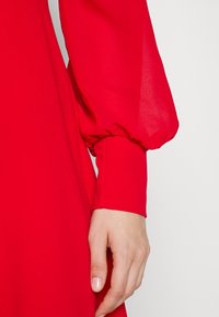 Punainen mekko, jonka hiha on kevyestä kankaasta ja puffimaisesta muotoilusta. Hihansuu on tiukka, korostaen sileää pintaa ja elävää väriä.