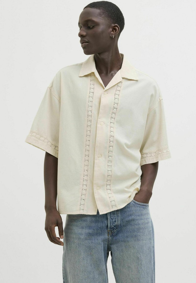 Camisa de botones de manga corta en color crema con detalles de ojales verticales, que presenta un cuello estructurado y un corte relajado, combinada con jeans de color azul claro.
