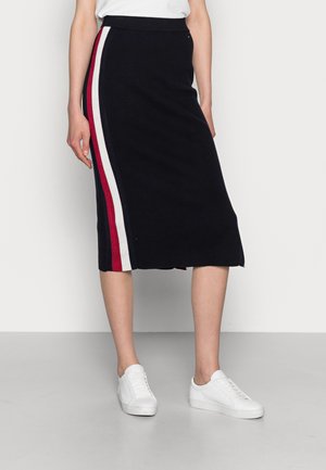 Vrouw draagt een zwarte midirok met verticale rode en witte strepen aan de zijkant, gecombineerd met witte sneakers en een witte top.