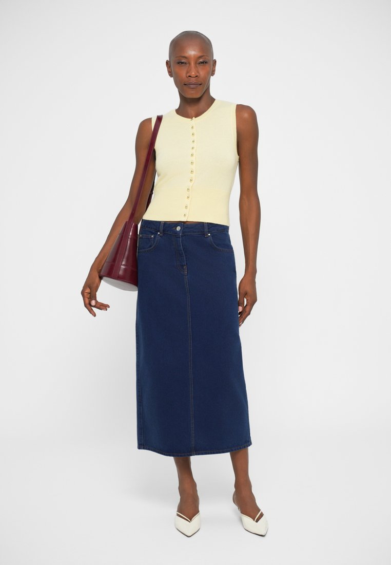 Haut sans manches crème avec des boutons, associé à une jupe maxi en denim bleu foncé et des chaussures blanches à bout pointu. Un sac à main bordeaux complète le look.