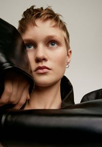 Chaqueta de cuero negra con un amplio cuello, enmarcando un rostro con cabello corto, piel clara y ojos azules, que presenta un pendiente de aro de plata.