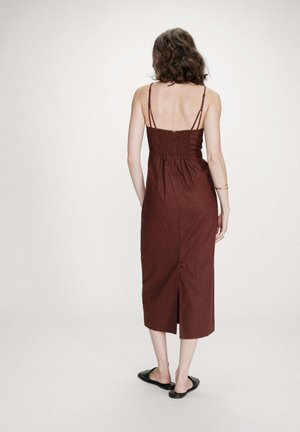 Femme debout de dos portant une robe midi marron sans manches à fines bretelles et des chaussures noires sans lacets sur un fond uni.