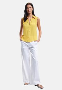 Yellow sleeveless blouse with two pocket flaps, paired with wide white trousers. Model wears flat black sandals with thin straps.