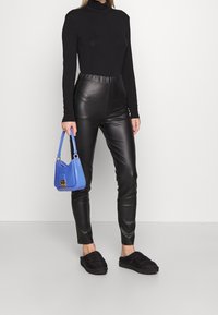 Svart polotröja, höga svarta byxor i konstläder, svarta vadderade tofflor, håller i en liten blå handväska med guldkant.