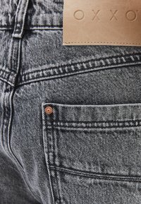Jeans de mezclilla negra con una superficie texturizada, que cuentan con una etiqueta de cuero con "OXXO" y un remache de cobre en el bolsillo trasero.