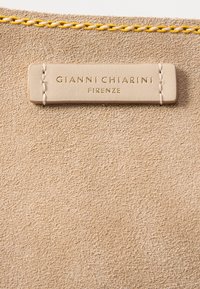 Tissu beige texturé avec une étiquette en cuir cousue indiquant "Gianni Chiarini Firenze" et un cordon torsadé jaune et blanc au-dessus.