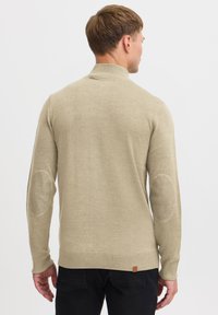 Jersey de punto beige con cuello alto, puños acanalados y parches en los codos. Textura suave, diseño ajustado y parche con el logo en el dobladillo.