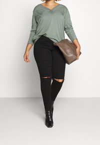 Top lungo a maniche nere in verde oliva con scollatura a V, abbinato a jeans skinny neri strappati e stivaletti neri, mentre tiene una borsa a tracolla marrone.