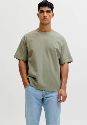 Mann mit kurzem dunklem Haar, der ein locker sitzendes hellgrünes Kurzarm-T-Shirt und hellblaue Jeans trägt und mit einer Hand in der Tasche steht.