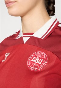 Piros mez fehér kiegészítőkkel, klasszikus gallérral és logóval. Simító anyagból készült, sportos kialakítással és részletes varrással.