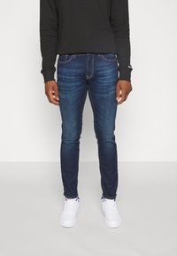 Man med slim fit mörkblå jeans, svart långärmad skjorta och vita sneakers som står mot en neutral ljus bakgrund.