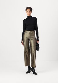 Sudadera negra de manga larga con cuello de tortuga, pantalones anchos metálicos dorados, botines negros de tacón, sosteniendo un pequeño bolso negro. Fondo minimalista.