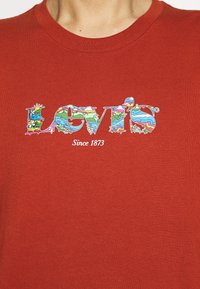 T-shirt en coton rouge présentant un logo graphique multicolore avec des paysages et des fleurs, avec les textes "Levi's" et "Since 1873" en dessous.