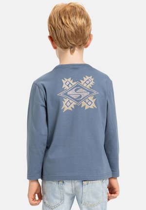 Long-sleeve blue shirt made from soft fabric, featuring a beige geometric design with an emblem on the back, worn with light denim jeans.