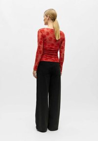 Sarkans apģērbs ar garām piedurknēm un rožu rakstu, ar savākto audumu, kombinācijā ar melnām biksēm ar platiem kāju galiem, skatīts no aizmugures.