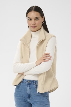 Vrouw met lang, donker haar die kleine creolen draagt, een crèmekleurig fleecevest over een witte coltrui en een blauwe spijkerbroek, staand met gekruiste armen.