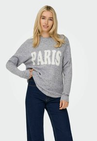 Un pull en tricot gris avec "PARIS" en blanc, des manches larges et une coupe décontractée. Associé à un pantalon en denim bleu foncé.
