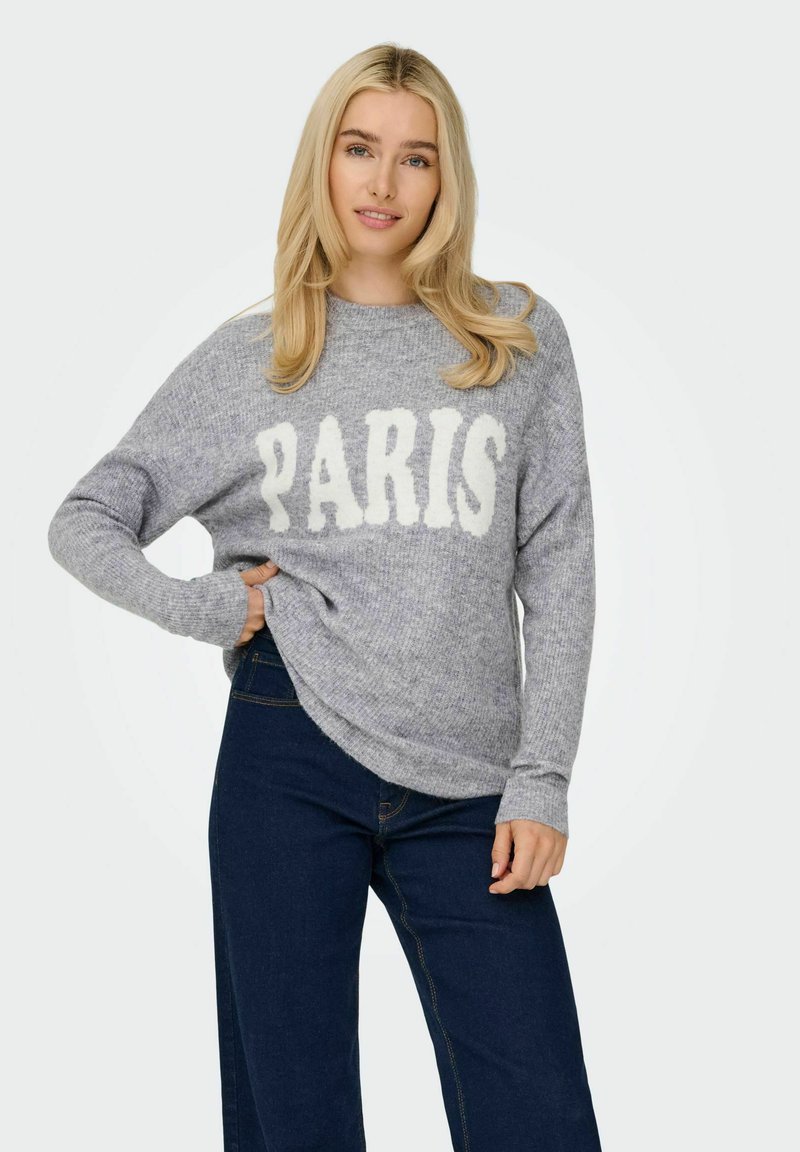 Un pull en tricot gris avec "PARIS" en blanc, des manches larges et une coupe décontractée. Associé à un pantalon en denim bleu foncé.