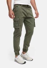 Homme portant un pantalon jogger cargo vert olive et des baskets blanches, debout avec les mains détendues le long du corps.