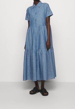 Kobieta ubrana w niebieską, krótki rękaw, zapinaną na guziki sukienkę midi z falbanami oraz czarne sandały z odkrytymi palcami na jednolitym tle.