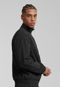 Svart zip-up jacka i lätt tyg, hög krage, rynkade ärmslut och elastisk midja, med en strukturerad finish.