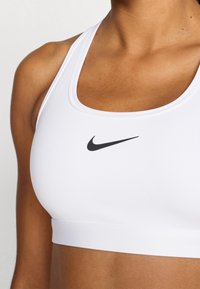 Λευκό αθλητικό στράπλες με τετράγωνο ντεκολτέ και λεπτές τιράντες. Διαθέτει μικρό μαύρο λογότυπο Nike στο μπροστινό μέρος. Ομαλή υφή υφάσματος.