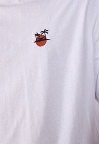 White T-shirt featuring a small, embroidered round design with two palm trees and a sunset in red and black. Soft cotton material.