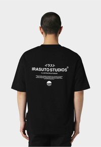 Fekete pamut póló, fehér grafikai nyomattal a hátsó részen, amely tartalmazza az "IRASUTO STUDIOS" szöveget és egy kör alakú dizájnelemet.