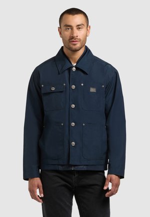 Moški, oblečen v temno modro srajco z gumbi, štirimi sprednjimi žepi in srebrnimi gumbi, stoji pred enobarvnim ozadjem.