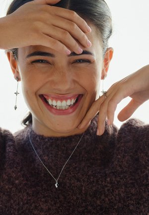 Collana d'argento e orecchini pendenti a forma di stella; la modella indossa un maglione scuro testurizzato, sorride con le mani sul viso.