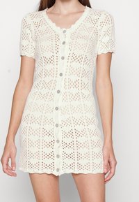 Abito bianco lavorato a maglia con motivo traforato, maniche corte, scollo smerlato e bottoni frontali. Tessuto strutturato e silhouette aderente.