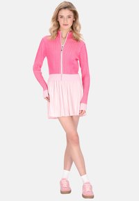 Rosa gerippter Reißverschluss-Pullover mit passendem plissiertem Rock. Stricktextur, kontrastierende hellrosa Bündchen und weiche rosa Sneakers mit weißen Socken.