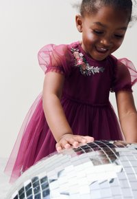 Lila tyllklänning med puffärmar, prydd med blomsterbroderier vid halsringningen. Barnet leker med en reflekterande discokula.